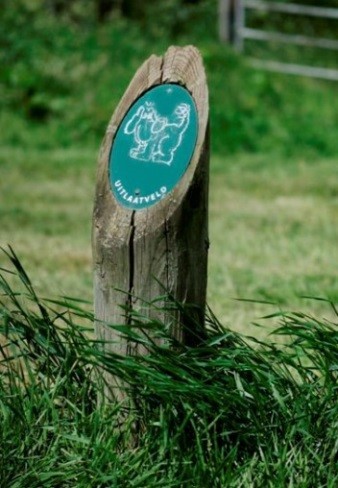 Een houten paaltje in het gras met daarop een groen plaatsen met daarop een afbeelding van een hond en de tekst: uitlaatveld.