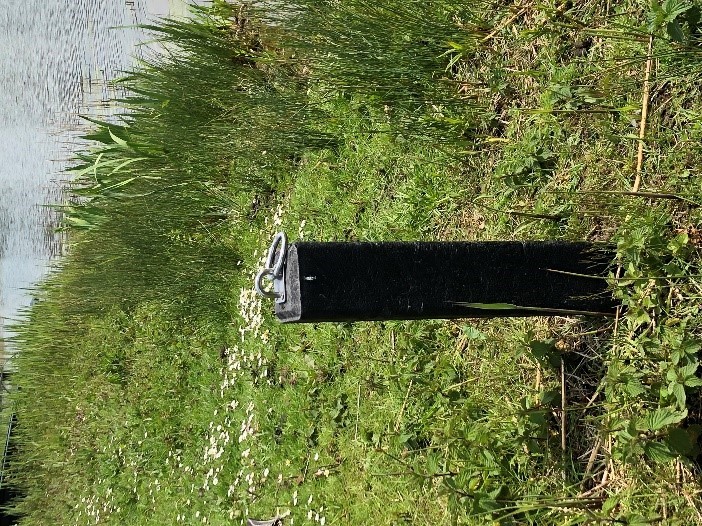 Een aanmeerpaal in het gras naast het water bij de Slottuin in Beuningen. Bovenop de paal zit een oog.