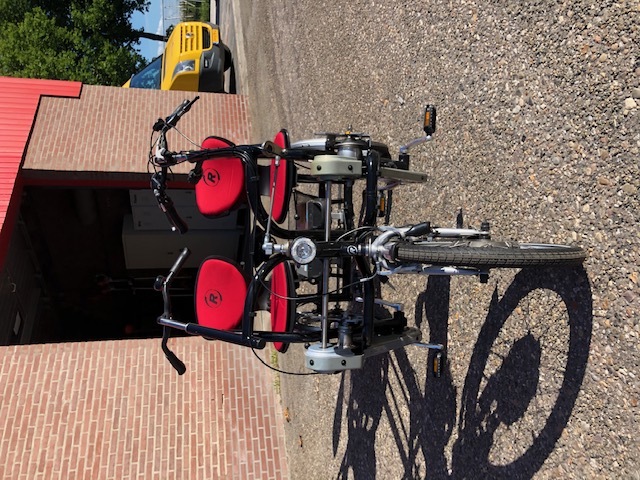 Een duofiets met rode zittingen, twee sturen en drie wielen staat voor een stenen loods geparkeerd.